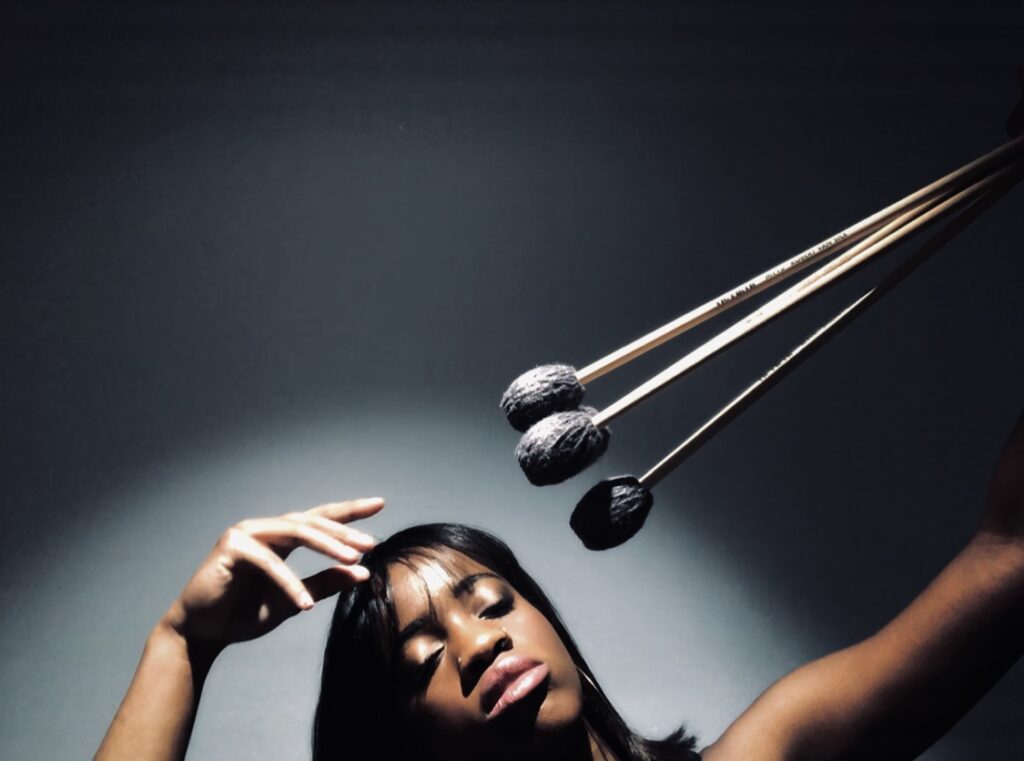 black female with long hair holding three percussion mallets above her head