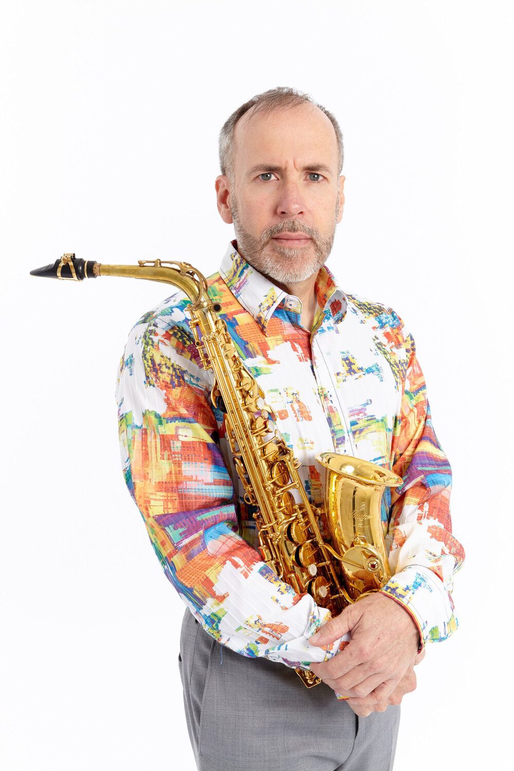 white male with grey short hair holding a brass saxophone in his arms and wearling a white and bright colors shirt