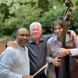 black male holding drum stick, white male with grey hair holding shoulders of friends, and white male with dark hair holding a double bass instument