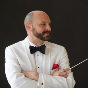 white male in white dinner jacket and black bow tie looking to his left smiling and holding conductor baton