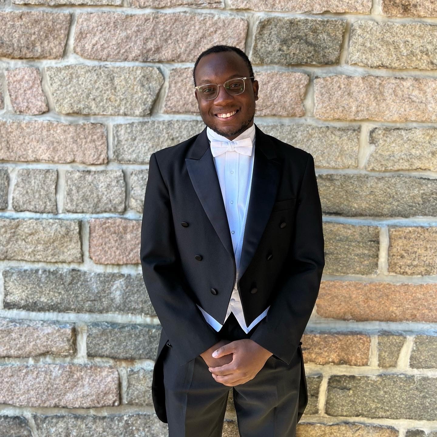 young black man wearing eye glasses and a formal tuxedo standing in front of stone wall