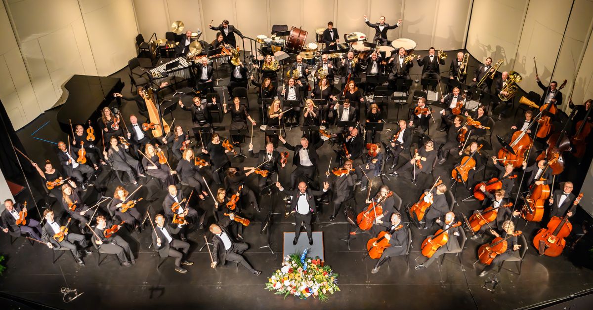 overhead photo of an orchestra on a stage waving to a camera high above the stage