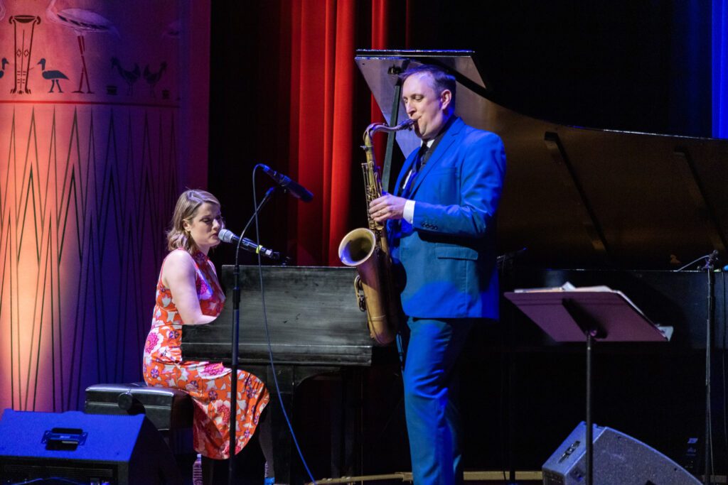 white female in pink dress playing a piano and singing with a tall white mael in a blue suite playing a saxphone