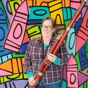 Amber Ferenz wearing a purple plaid shirt, holding their bassoon, in front of a colorful mural depicting crayons of all colors and sizes.