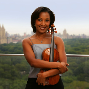 Kelly Hall-Tompkins wearing a silver-gray tank top and black pants, holding her violin in front of a distant city skyline and trees.