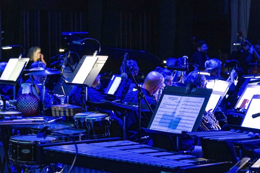 blue lighted stage featuring percussion instruments and orchestra members, lights on music stands