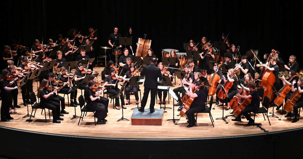 group of orchestra players made up of teenager males and females on a stage holding instruments led by black male music conductor. stage is light wood, background is black