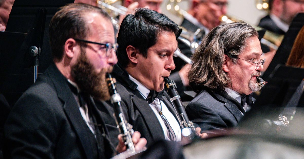 three white male musicians in tuxedos playing clarinet instruments in an orchestra