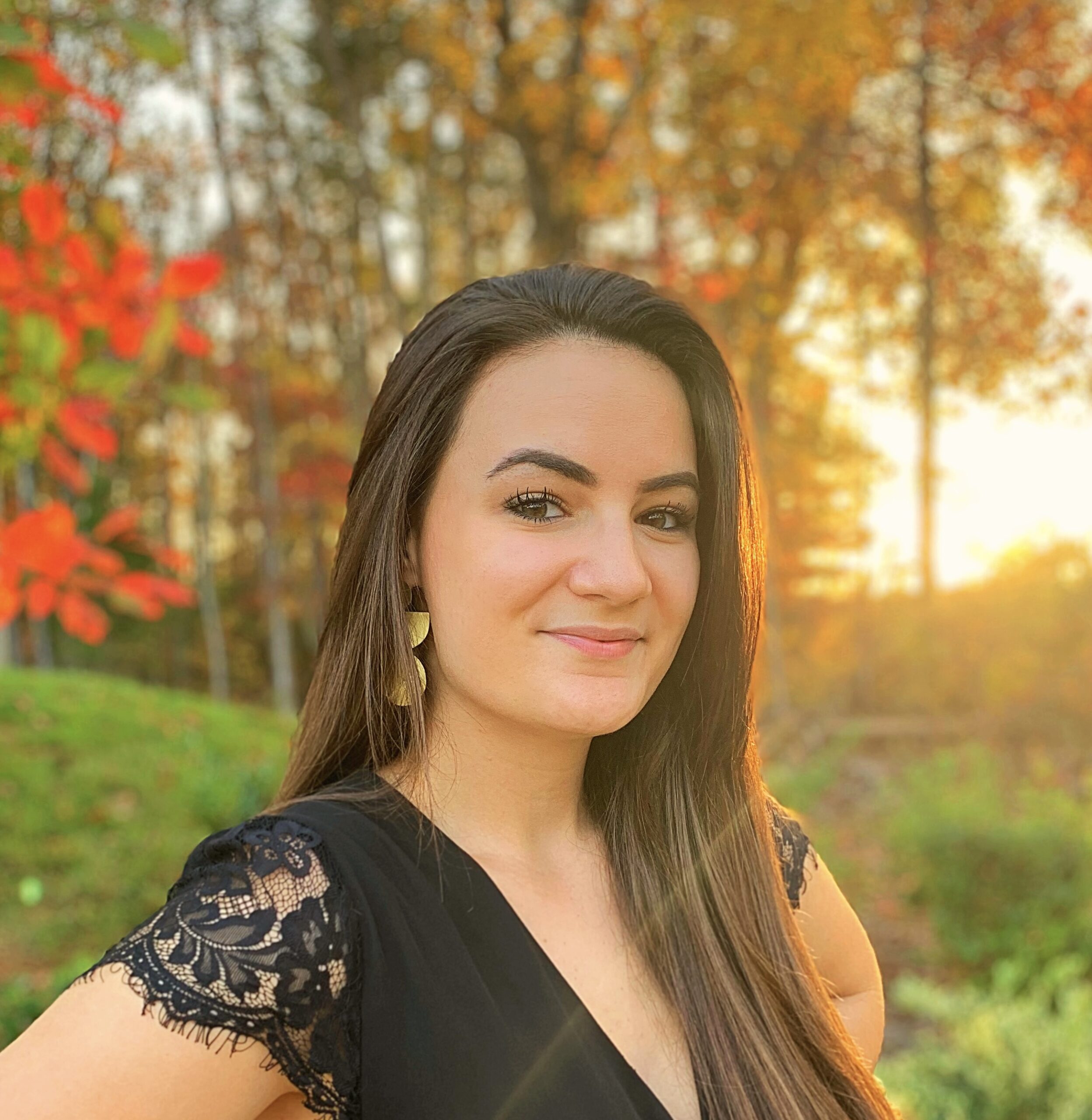 white female and black dress top withdark brown long hard looking at the camera standing in front of sunset and autumn leaves