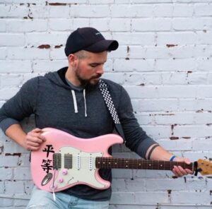 white man with a black goateee wearing a black baseball hat and grey sweatshirt holding a pink Fender electric guitar standing in front of a white painted brick wall