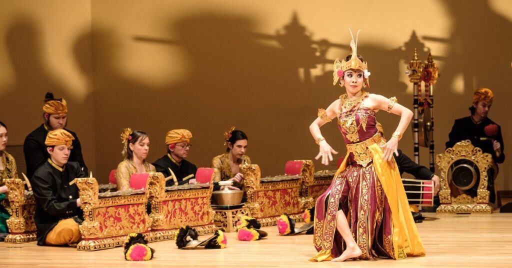 Asian female in gold and red Balinese style traditional costume dancing in front of five white musicians in traditional gold turban and white shirts playing gamelan metal percussions instruments