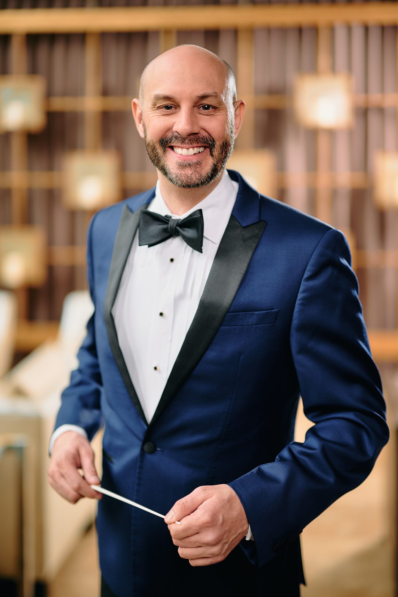 white male with goatee and bald head wearing a dark blue silk tuxedo with white shite and bowtie, smiling at cameras and holding a conductors baton