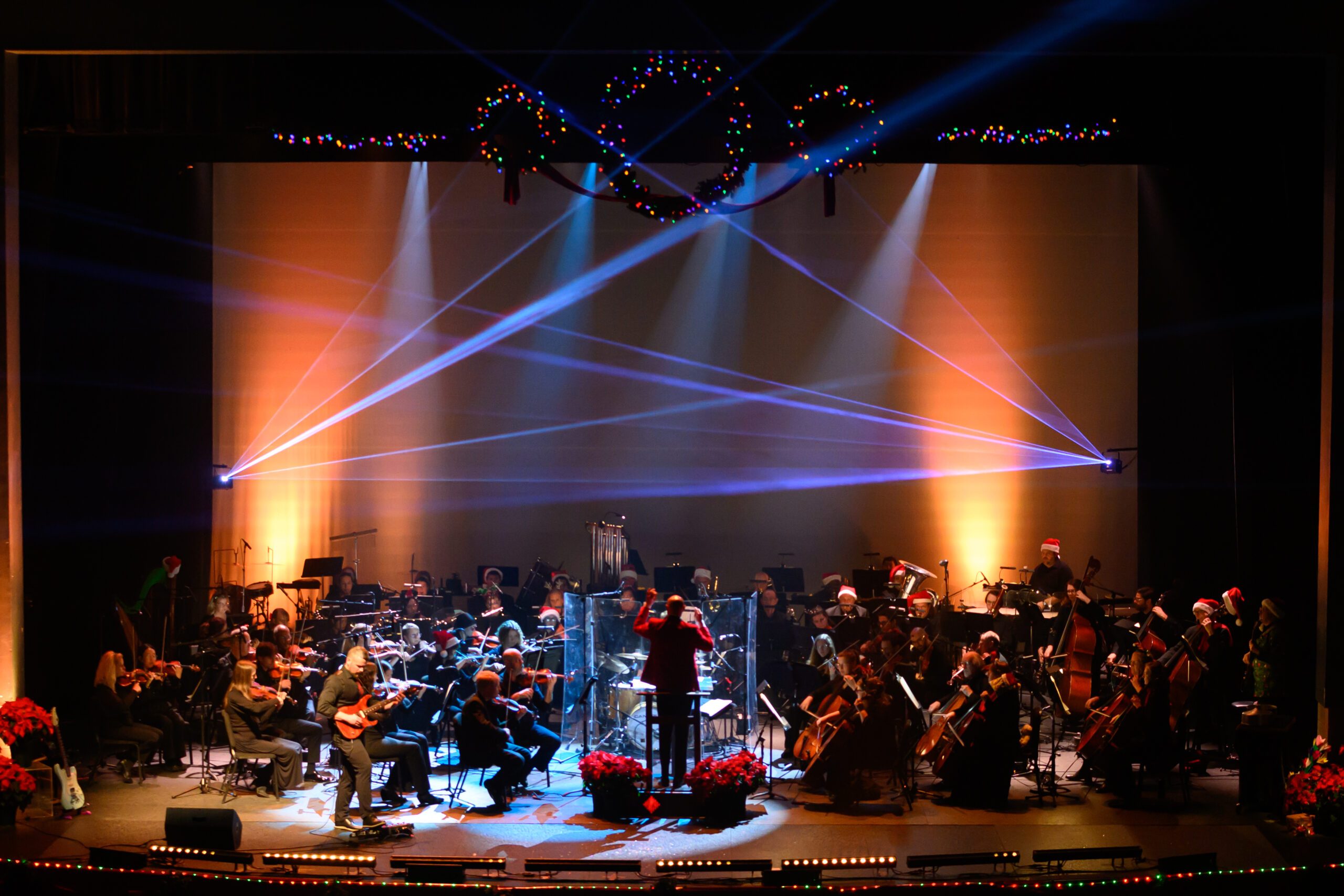 stage with an orchestra under flue and red lights with conductore in red coat and man playing elextric guitar