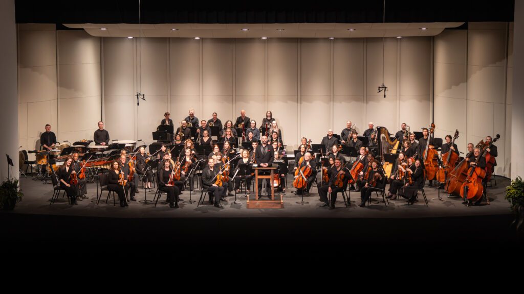 Group of orchestra musicians standing on a stage holding instruments and wearing black clothing smiling at a camera