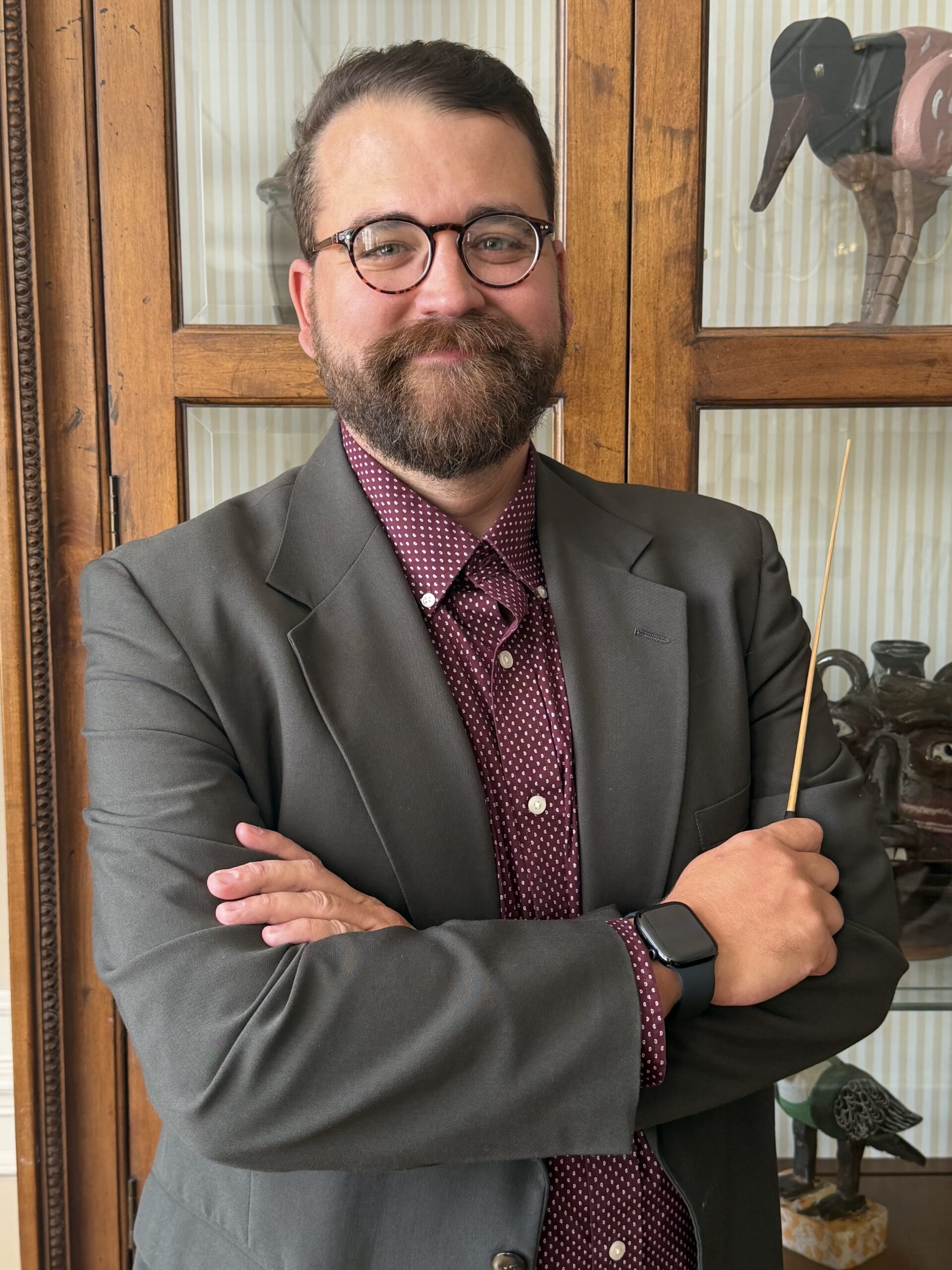 white male with brown hair and brown beard weearing horn rimmed eyeglasses smilling at the camera holding a conductor's baton and wearing a maroon shirt and grey suit coat